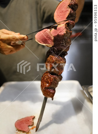 Picanha is a BBQ Steak meat grilled in charcoal. Knife cutting on skewer. Brazilian meat in a Churrascaria restaurant Picanha is a BBQ Steak meat grilled in charcoal. Knife cutting on skewer. Brazilian meat in a Churrascaria restaurant 95996313