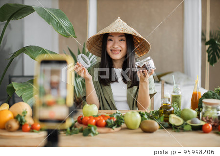 Beautiful smiling asian young woman blogger in traditional conical hat cooking organic healthy food Beautiful smiling asian young woman blogger in traditional conical hat cooking organic healthy food 95998206