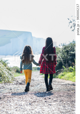 Two little girls in red checked dress and yellow trousers walking holding hands towards Cuckmere beach located between Seaford and Eastbourne in East Sussex, England. Seven sisters park. 96000237