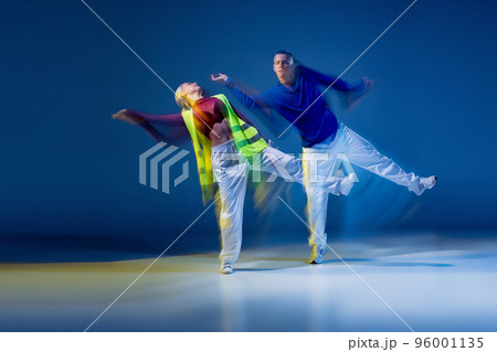 Portrait of young man and woman dancing isolated over dark blue background with mixed lights. Synchronic 96001135