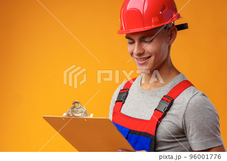 Young construction worker wearing hardhat and holding a clipboard on yellow background in studio 96001776
