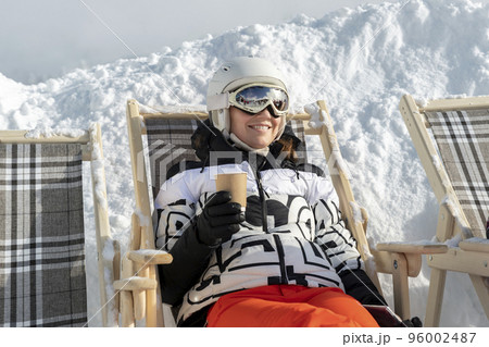 Portrait young adult happy smiling womah enjoy having fun drinking hot coffee tea sit in deckchair lounge area at alpine mountain european ski resort sunny cold winter day. Sunbathing outdoors 96002487