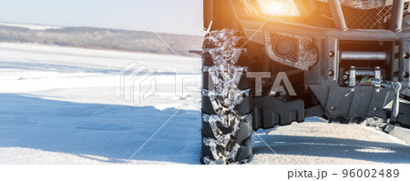 Front POV close-up detail view of quad bike offroad vehicle parked in snowdrift track on sunny snowy cold winter morning against clear blue sky. ATV adventure extreme sport. Nature country tour drive 96002489