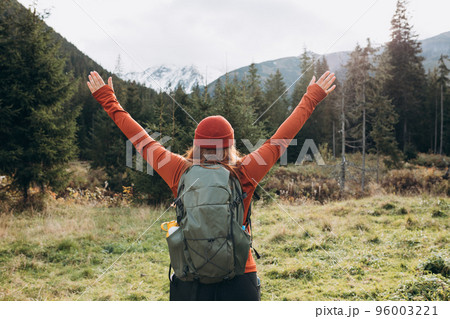 Back view of stylish woman with arms up, wearing green backpack and red hat, looking at mountain view while relaxing in nature. Travel and wanderlust concept. Successful woman enjoying triumph Back view of stylish woman with arms up, wearing green backpack and red hat, looking at mountain view while relaxing in nature. Travel and wanderlust concept. Successful woman enjoying triumph 96003221
