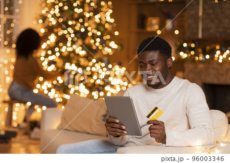 Glad millennial black man with tablet and credit card enjoy online shopping, wife decorates Christmas tree 96003346