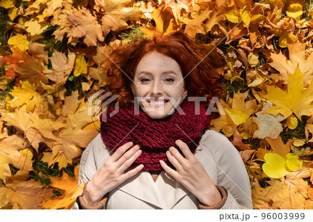 Smiling caucasian millennial woman with red hair in raincoat with scarf lies on ground with yellow leaves 96003999