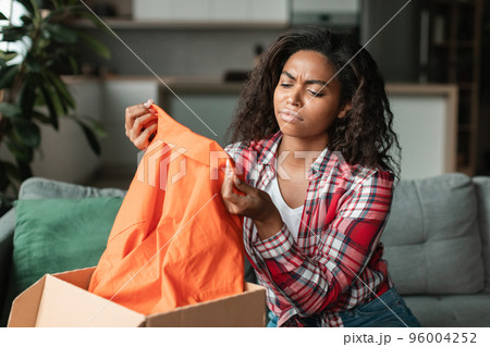 Sad dissatisfied black young female in casual looks at clothes, unpacks parcel in living room interior 96004252