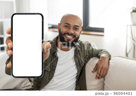 Happy latin man showing smartphone with white empty screen in hand, sitting on couch at home, mock up 96004401