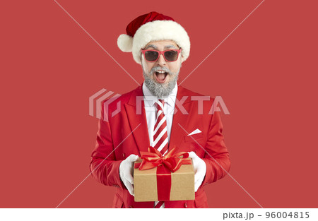 Old grey-haired happy showman in Santa style is giving a present, gift box on red background. 96004815