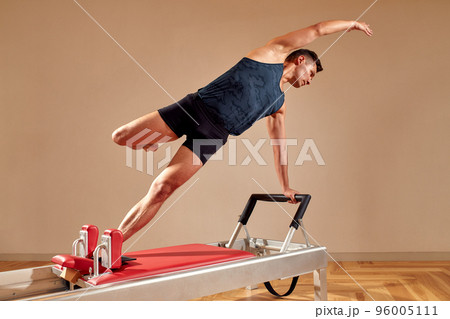 Side view of barefoot male athlete lying on pilates reformer and performing abs exercise during fitness workout. Pilates man concept. Side view of barefoot male athlete lying on pilates reformer and performing abs exercise during fitness workout. Pilates man concept. 96005111
