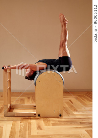 Handsome pilates male instructor performing stretching balance fitness exercise on small barrel equipment, at the pilates studio modern onterior indoor. 96005112