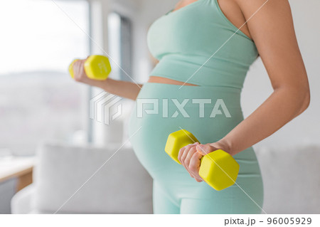 Pregnant woman prenatal training at home doing fitness strength training exercise with dumbbell weights. Body workout arm curls closeup of belly and arms. Easy workout in second and third timester Pregnant woman prenatal training at home doing fitness strength training exercise with dumbbell weights. Body workout arm curls closeup of belly and arms. Easy workout in second and third timester 96005929