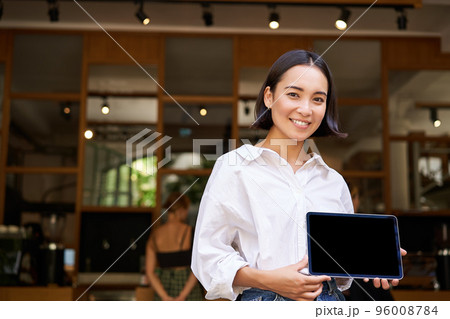 Smiling asian female manager, cafe owner, showing tablet screen, demonstrating smth on her device 96008784