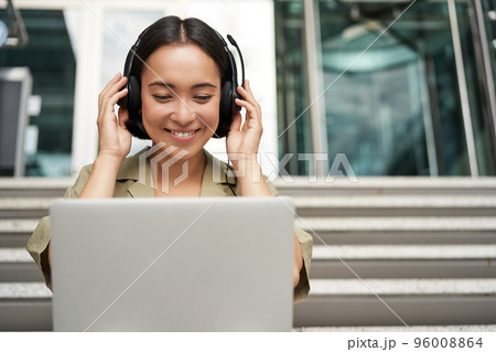 Portrait of young asian woman sitting with laptop and headphones, watching video, does online course on computer, sitting on stairs outdoors 96008864