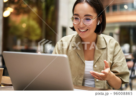 Online meeting. Young urban girl, asian woman talking with laptop, video chat, gesturing, sitting in an outdoor cafe and working remotely 96008865