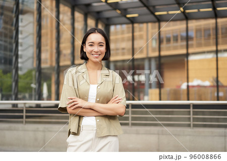Portrait of beautiful young woman in casual clothes, smiling, posing outdoors on an empty street near glass building 96008866