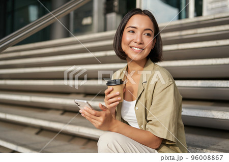 Happy urban girl drinks her takeaway coffee and scrolls feed on smartphone. Asian woman sits on stairs with tea and holding mobile phone 96008867