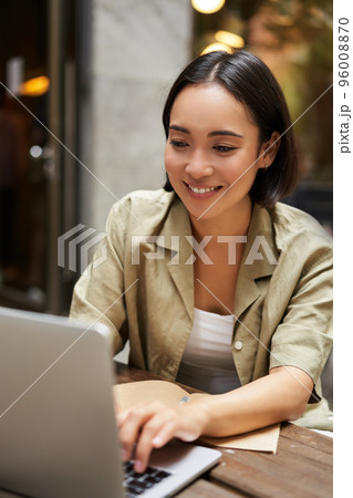 Vertical shot of young asian woman working on remote from outdoor cafe, sitting with laptop and smiling, studying 96008870
