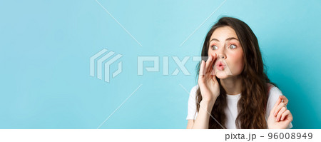 Cute glamour gossip girl spreading rumours, whispering news to someone, standing voer blue background Cute glamour gossip girl spreading rumours, whispering news to someone, standing voer blue background 96008949
