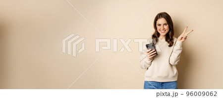 Beautiful smiling woman showing peace sign, holding mobile phone, standing over beige background Beautiful smiling woman showing peace sign, holding mobile phone, standing over beige background 96009062
