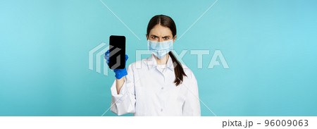 Angry and confused woman doctor in face mask, gloves, showing smartphone app, mobile screen, frowning upset, standing over blue background 96009063
