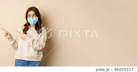 Health and covid-19 concept. Cute brunette woman in medical face mask, pointing fingers left at logo, banner, showing copy space, standing over beige background Health and covid-19 concept. Cute brunette woman in medical face mask, pointing fingers left at logo, banner, showing copy space, standing over beige background 96009317