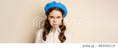 Close up portrait of young beautiful woman sulking, looking angry and disappointed, grimacing displeased, standing over beige background 96009319