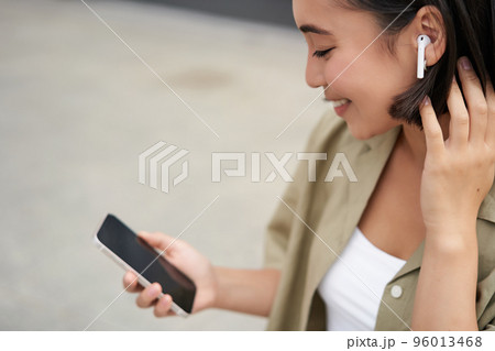 Close up portrait of asian girl, looking at mobile screen, listening music in headphones. Woman with earphones walks on street 96013468