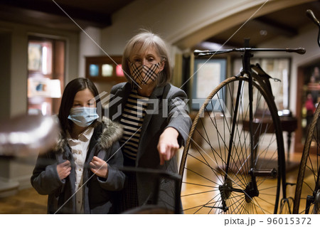 Tween schoolgirl and elderly female tutor in face masks viewing vintage bicycle in museum 96015372