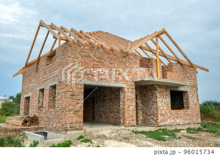 Building site with unfinished brick house with wooden roofing frame for future roof under construction. Building site with unfinished brick house with wooden roofing frame for future roof under construction. 96015734