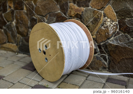 Big roll of white industrial electrical cable on large wooden reel outdoors on sunny pavement on stone fence background. Professional construction site cable, reliable wiring and durability. Big roll of white industrial electrical cable on large wooden reel outdoors on sunny pavement on stone fence background. Professional construction site cable, reliable wiring and durability. 96016140