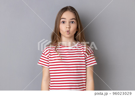 Funny surprised little girl wearing striped T-shirt making fish face with pout lips and looking amazed, choking with idiotic comical silly expression. Indoor studio shot isolated on gray background. 96023029