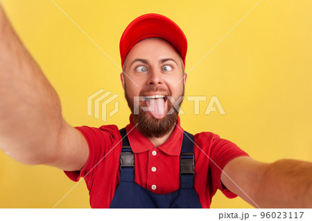 Funny handyman wearing blue uniform taking selfie, looking at camera with crossed eyes and showing tongue out POV, point of view of photo. Indoor studio shot isolated on yellow background. Funny handyman wearing blue uniform taking selfie, looking at camera with crossed eyes and showing tongue out POV, point of view of photo. Indoor studio shot isolated on yellow background. 96023117