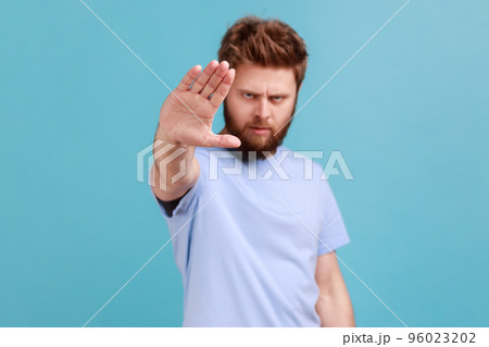 Portrait of strict bearded man showing stop sign at camera, standing with frowning face and bossy expression, meaning caution to avoid danger or mistake. Indoor studio shot isolated on blue background 96023202