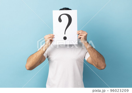 Portrait of man wearing white T-shirt hiding his face behind white paper with question mark, finding smart solution, asking for advice. Indoor studio shot isolated on blue background. Portrait of man wearing white T-shirt hiding his face behind white paper with question mark, finding smart solution, asking for advice. Indoor studio shot isolated on blue background. 96023209