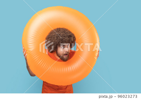 Portrait of man with astonished facial expression looking trough bright orange rubber ring away with open mouth, surprised by vacation. Indoor studio shot isolated on blue background. Portrait of man with astonished facial expression looking trough bright orange rubber ring away with open mouth, surprised by vacation. Indoor studio shot isolated on blue background. 96023273