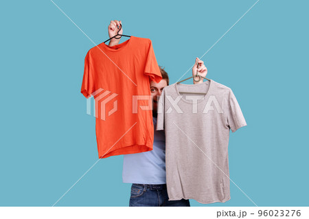 Portrait of optimistic young adult handsome bearded man peeping from T-shorts on hangers, choosing attire, expressing positive emotions. Indoor studio shot isolated on blue background. Portrait of optimistic young adult handsome bearded man peeping from T-shorts on hangers, choosing attire, expressing positive emotions. Indoor studio shot isolated on blue background. 96023276