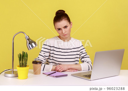 Bored unhappy woman office manager wearing striped shirt sitting at workplace looking at camera, tired of online conference. Indoor studio studio shot isolated on yellow background. 96023288