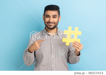 Viral hashtag and successful blogging. Smiling businessman with beard pointing at large yellow hash symbol, wearing striped shirt. Indoor studio shot isolated on blue background. Viral hashtag and successful blogging. Smiling businessman with beard pointing at large yellow hash symbol, wearing striped shirt. Indoor studio shot isolated on blue background. 96023433