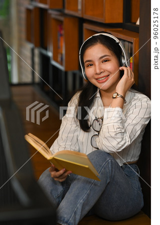 Cheerful Asian female student listening to music on headphone and reading book in college library 96025178