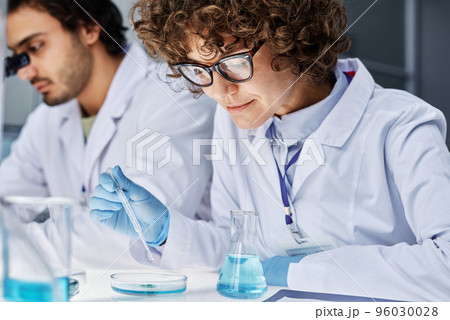 Young clinician adding some drops of fluid in pipette into petri dish with bacteria while carrying out scientific experiment with colleague 96030028