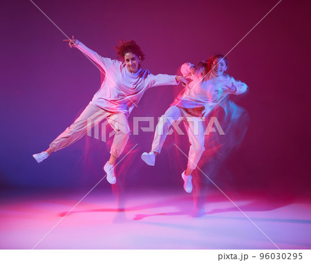 Portrait of two young girls dancing hip-hop isolated over gradient violet background in neon with mixed light. Jumping high 96030295