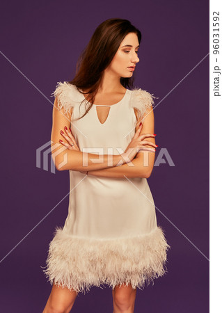 Beautiful brunette woman in elegant cocktail white dress is posing at studio on purple background. 96031592