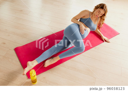 Portrait of young pregnant woman in sportswear lying on mat and holding belly, stretching exercises indoors on a daytime 96038123