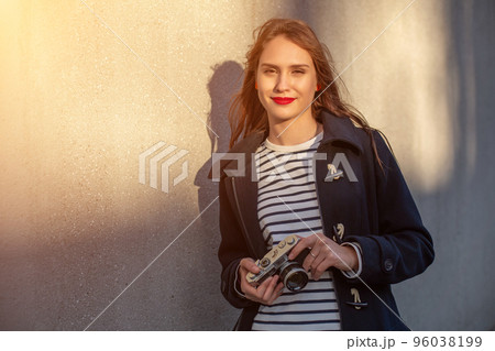 Smiling female photographer in jacket standing in front of wall ready to make new photo. Sun flare 96038199