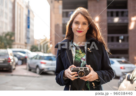Outdoor autumn smiling lifestyle portrait of pretty young woman, having fun in the city with camera, travel photo of photographer. 96038440