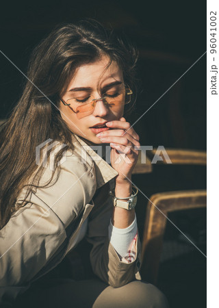 beautiful brunette girl in a sunglasses dressed in white blouse and beige coat posing in a chair. Stylish trendy fashion outlook 96041002