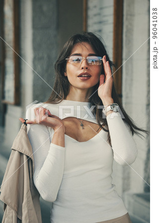 beautiful brunette girl in a sunglasses dressed in white blouse with a beige coat in hands. Stylish trendy fashion outlook beautiful brunette girl in a sunglasses dressed in white blouse with a beige coat in hands. Stylish trendy fashion outlook 96041038