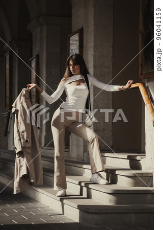 beautiful brunette girl dressed in white blouse, pants, sneakers with a handbag and camel coat in hands. Stylish trendy fashion outlook 96041159