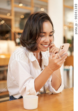 Vertical shot of brunette asian girl sitting in cafe, using smartphone app, chatting on mobile phone and smiling Vertical shot of brunette asian girl sitting in cafe, using smartphone app, chatting on mobile phone and smiling 96041389
